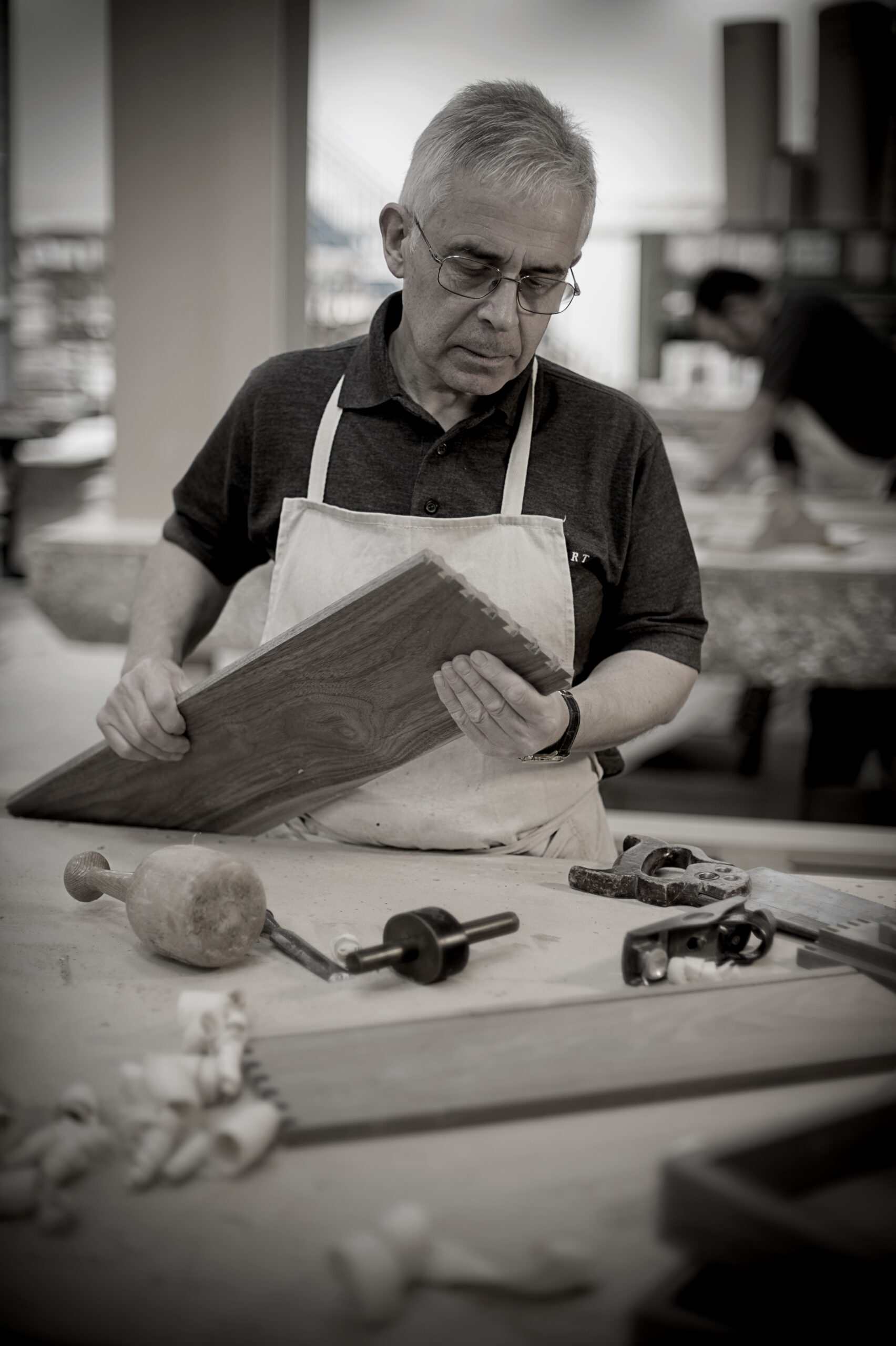 A man examining a piece of wood, surrounded by woodworking tools on a table